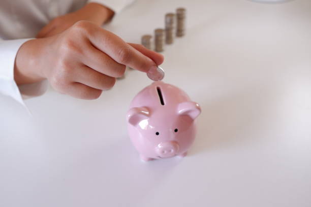 Businessperson Inserting Coins In Piggy Bank With Stack Of Coins Over The Desk